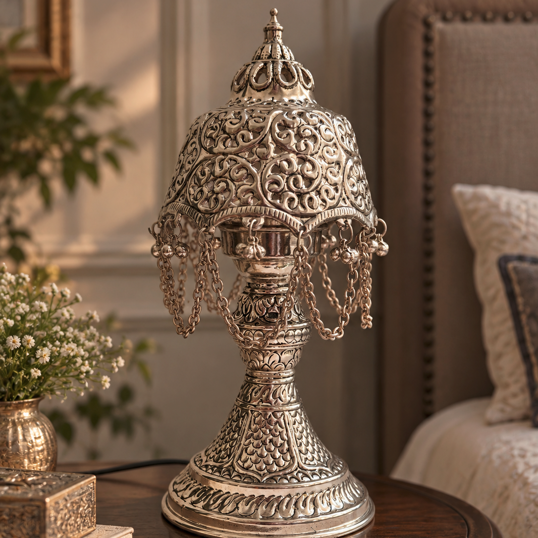 An ornate silver-toned metal table lamp with intricate filigree detailing and hanging chains, displayed on a dark wooden side table in a cozy bedroom setting.