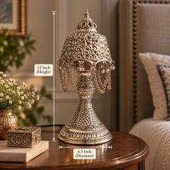 An ornate silver-toned metal table lamp with intricate filigree detailing and hanging chains, shown on a wooden table in a furnished room. White measurement lines indicate a height of 12 inches and a base diameter of 6.
