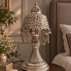 An ornate silver-toned metal table lamp with intricate filigree detailing and hanging chains, displayed on a dark wooden side table in a cozy bedroom setting.