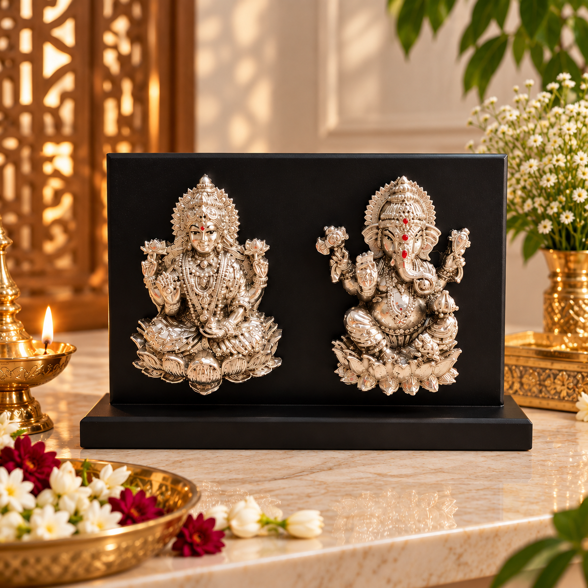 A decorative black rectangular plaque or box on a marble surface, featuring two ornate silver-toned deity sculptures facing each other. The scene is styled with a brass oil lamp, flowers, and greenery in a warm indoor setting.