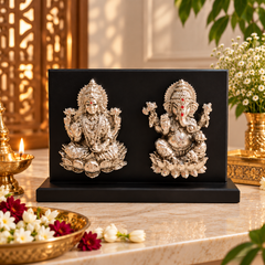 A decorative black rectangular plaque or box on a marble surface, featuring two ornate silver-toned deity sculptures facing each other. The scene is styled with a brass oil lamp, flowers, and greenery in a warm indoor setting.