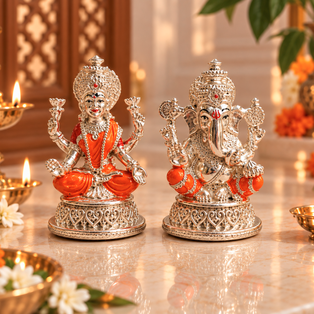 Two ornate silver deity statues, one of Ganesha and one of Hanuman, decorated with bright orange fabric and detailed with rhinestones, displayed on a polished marble surface with lit oil lamps in the background.