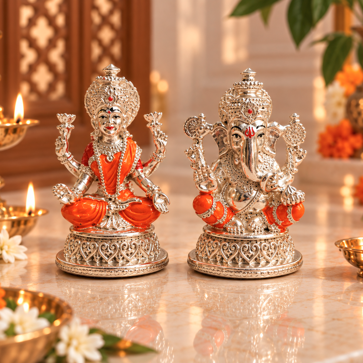 Two ornate silver deity statues, one of Ganesha and one of Hanuman, decorated with bright orange fabric and detailed with rhinestones, displayed on a polished marble surface with lit oil lamps in the background.