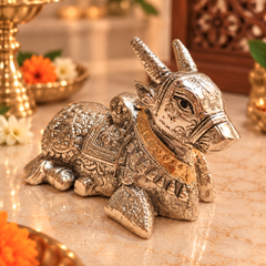 An ornate silver-toned metallic cow figurine with intricate engraved patterns and a gold-colored collar, displayed on a polished marble surface with warm decorative background elements.