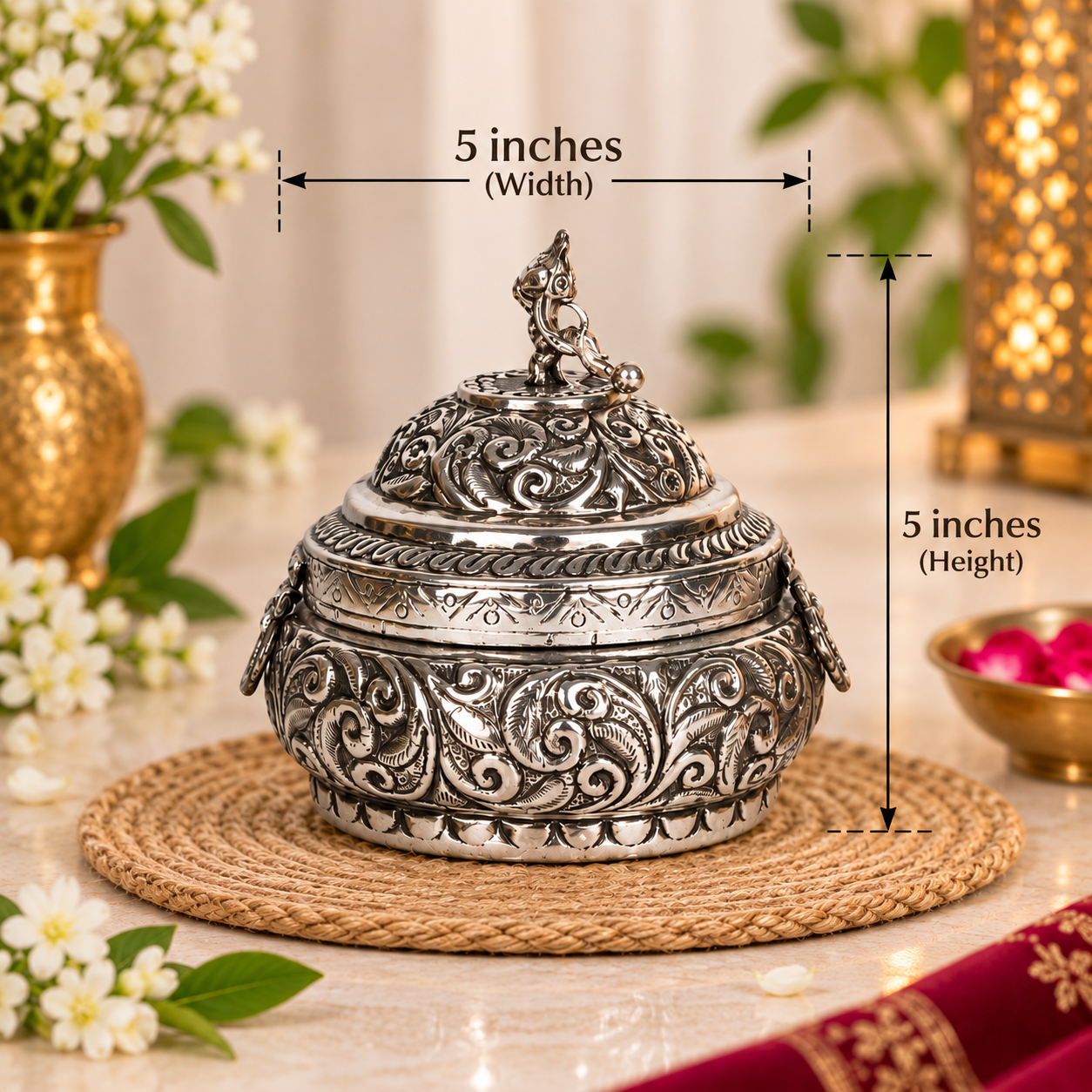 An ornate silver-toned metal lidded container with embossed scrollwork, shown on a woven placemat with overlaid dimension lines indicating 5 inches width and 5 inches height. The scene is styled with flowers, greenery, and warm ambient decor in the background.