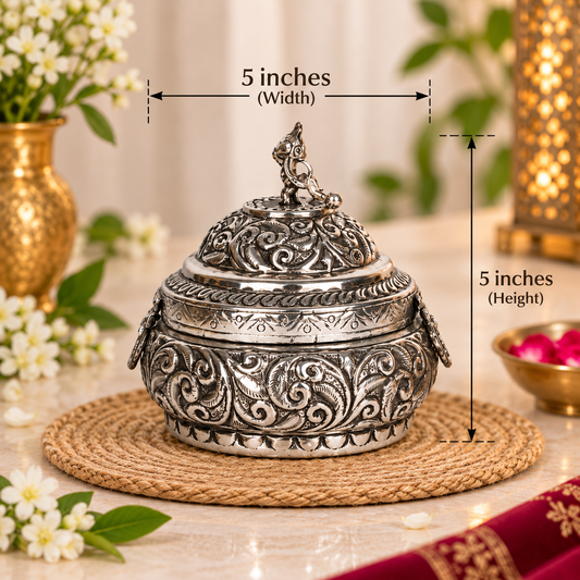 An ornate silver-toned metal lidded container with embossed scrollwork, shown on a woven placemat with overlaid dimension lines indicating 5 inches width and 5 inches height. The scene is styled with flowers, greenery, and warm ambient decor in the background.