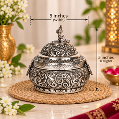 An ornate silver-toned metal lidded container with embossed scrollwork, shown on a woven placemat with overlaid dimension lines indicating 5 inches width and 5 inches height. The scene is styled with flowers, greenery, and warm ambient decor in the background.