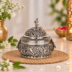 An ornate silver-toned metal lidded container with intricate embossed scrollwork, displayed on a woven placemat with decorative flowers and a warm lantern in the background.
