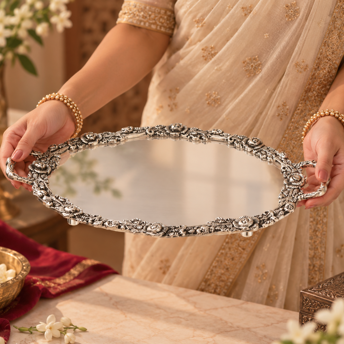 An ornate oval silver mirror with intricate floral and scrollwork handles, held by a person wearing a beige and gold sari. The background features a warm, decorated interior with flowers and a marble table.