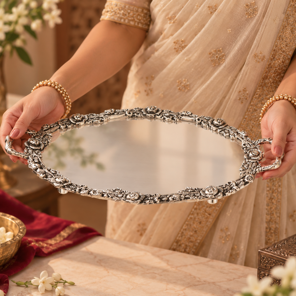 An ornate oval silver mirror with intricate floral and scrollwork handles, held by a person wearing a beige and gold sari. The background features a warm, decorated interior with flowers and a marble table.
