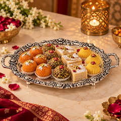An ornate silver serving tray filled with assorted Indian sweets and desserts, including laddoo, chikoo, and square sweets, arranged on a decorated table with flowers, candles, and a red fabric cloth.