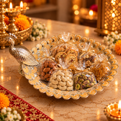 A decorative silver peacock-shaped serving tray filled with assorted packaged snacks, cookies, almonds, cashews, and pistachios, set on a warm lit table with candles and floral decor in the background.