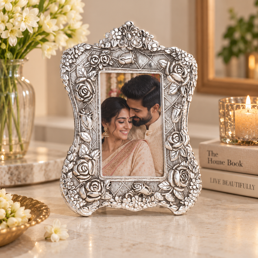 An ornate silver-toned rectangular photo frame with embossed floral and scrollwork details, standing on a marble surface. The frame contains a photo of a smiling couple, with a vase of white flowers, a lit candle on books, and a mirror in the softly lit background.