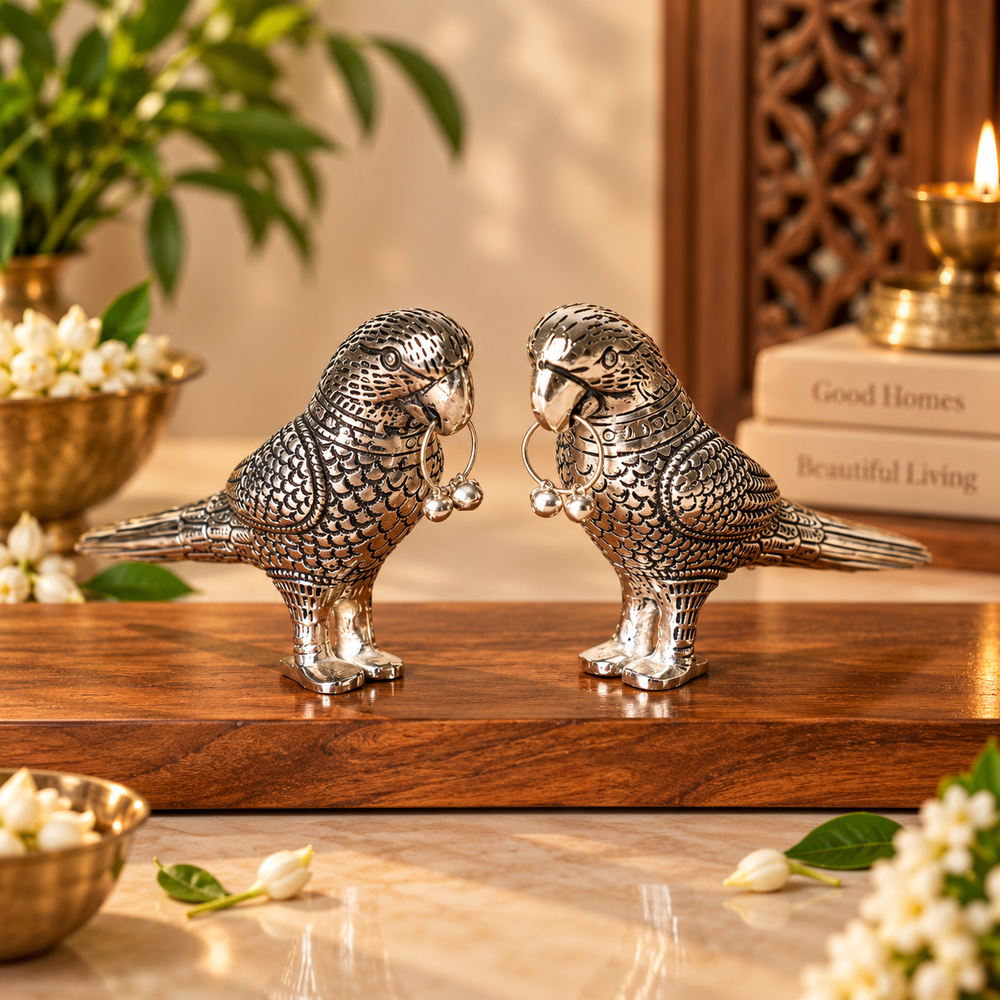 Two ornate silver bird figurines with detailed feather engravings and hanging bells, displayed on a wooden surface in a warm, styled interior setting.