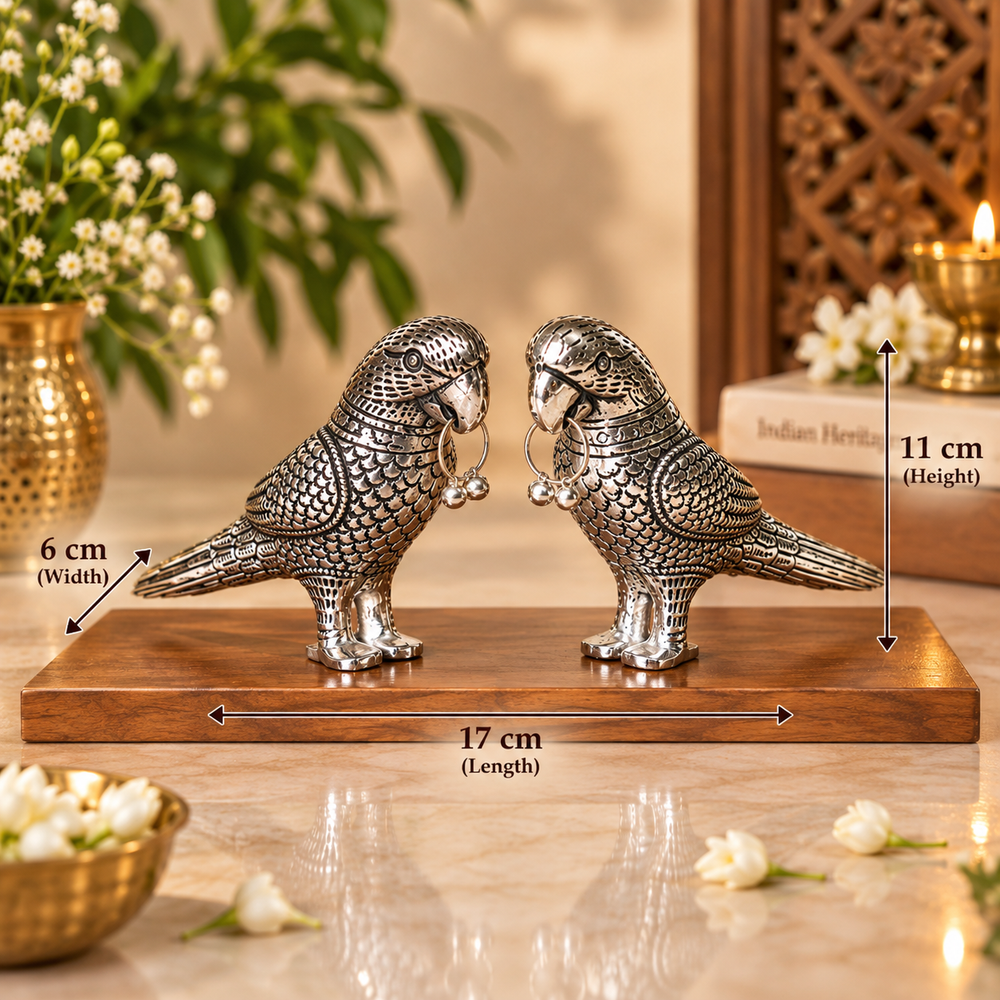 A pair of silver bird figurines standing on a wooden base, shown with overlaid dimension arrows and labels indicating width 17 cm, height 11 cm, and depth 6 cm. The scene is styled indoors with flowers, a candle, and decorative objects in the background.