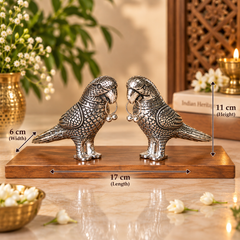 A pair of silver bird figurines standing on a wooden base, shown with overlaid dimension arrows and labels indicating width 17 cm, height 11 cm, and depth 6 cm. The scene is styled indoors with flowers, a candle, and decorative objects in the background.