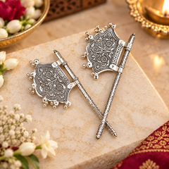 Two ornate silver-toned decorative axe-shaped accessories with intricate engraved patterns and small bead accents, displayed crossed on a beige stone slab with floral props and warm ambient lighting.