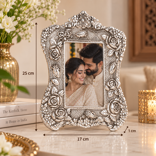 An ornate silver-toned metal picture frame with embossed floral details, shown standing on a tabletop in a styled indoor setting. Black dimension lines and text indicate the frame is approximately 25 cm tall, 17 cm wide, and has a 1 cm bevel.