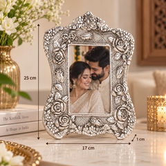 An ornate silver-toned metal picture frame with embossed floral details, shown standing on a tabletop in a styled indoor setting. Black dimension lines and text indicate the frame is approximately 25 cm tall, 17 cm wide, and has a 1 cm bevel.