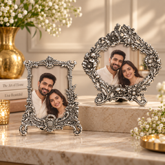 Two ornate silver-toned metal picture frames with floral scrollwork, each holding a portrait of a couple, displayed on a beige marble surface with decorative flowers and candles in a warm indoor setting.