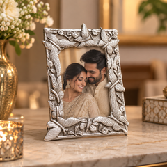 An ornate silver-toned rectangular picture frame with carved floral and leaf motifs, standing on a marble surface in a warmly lit indoor setting. The frame contains a photo of a smiling couple, and decorative flowers and a candle are visible in the background.