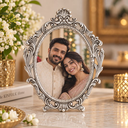 An ornate oval silver picture frame with intricate scrollwork and a small stand, holding a portrait of a smiling couple. The frame is displayed on a marble table with decorative flowers, books, and gold-toned candle holders in a warm, festive indoor setting.