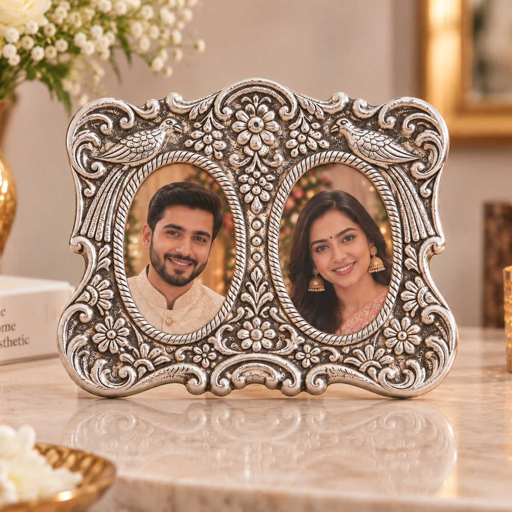 An ornate silver-toned double oval photo frame with intricate floral and scrollwork details, displayed on a marble surface in a warmly lit home setting with flowers and candles in the background.