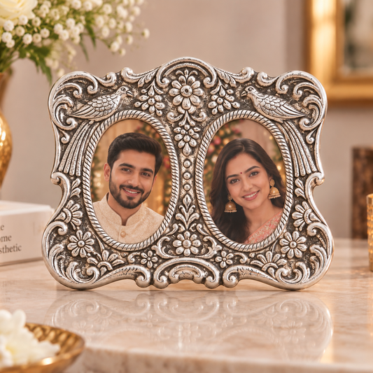 An ornate silver-toned double oval photo frame with intricate floral and scrollwork details, displayed on a marble surface in a warmly lit home setting with flowers and candles in the background.