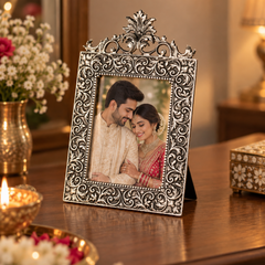 An ornate silver-toned metal picture frame with intricate scrollwork and a decorative crest at the top, holding a photo of a couple. The frame is displayed on a polished wooden table in a warmly lit room with flowers and candles in the background.
