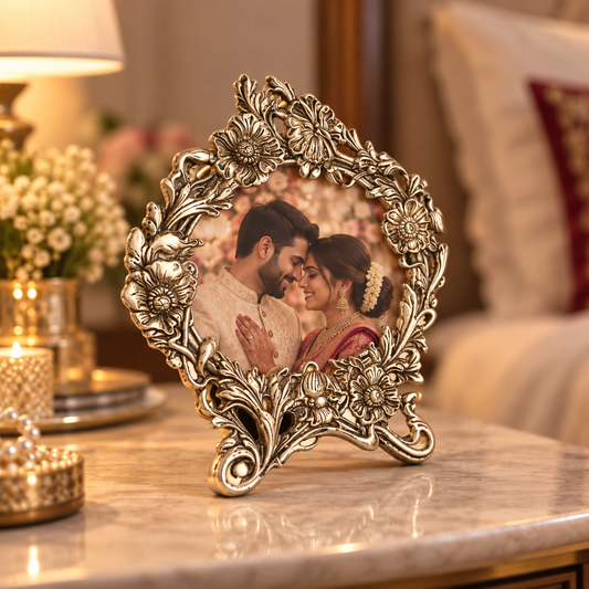 An ornate vintage-style metallic picture frame with floral detailing, standing on a marble table in a warmly lit living room. The frame contains a printed photo of a couple.