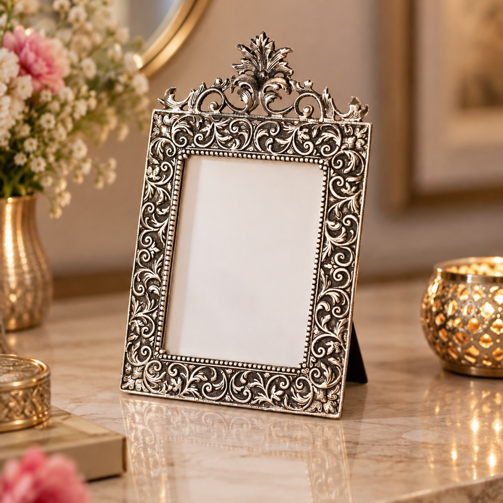 An ornate silver-toned metal picture frame with intricate scrollwork and a decorative crest at the top, displayed on a marble surface with flowers and gold-toned decor in the background.