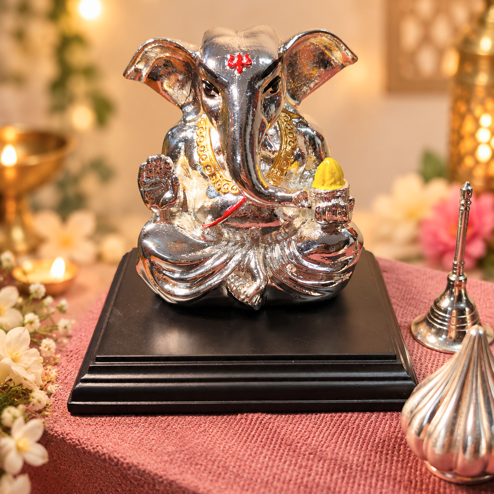 A metallic silver Ganesha statue with gold and red accents, seated on a black rectangular base. The statue is decorated with a red tilak on the forehead and holds a yellow modak in one hand.