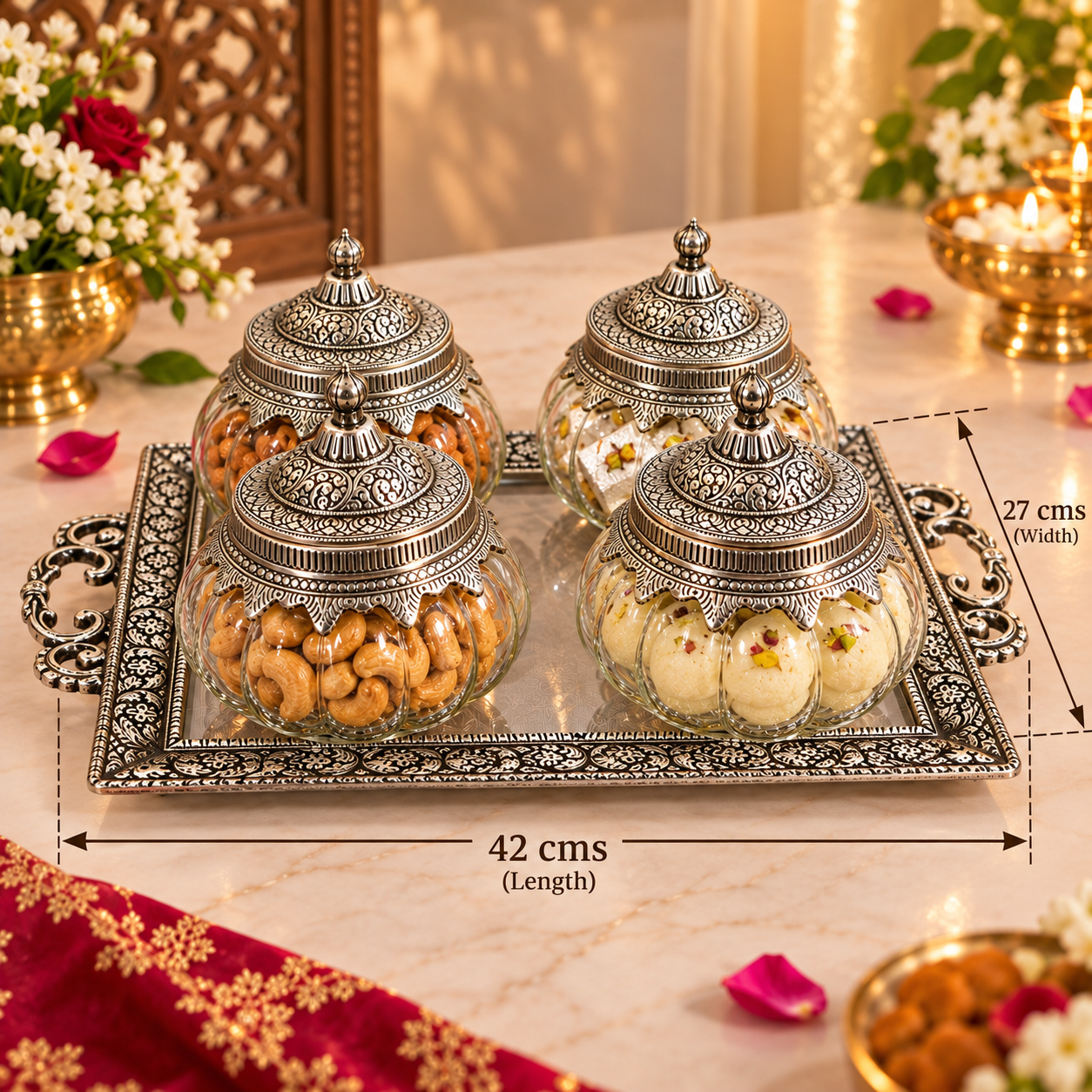 Four ornate silver-toned glass jars with matching lids are arranged on a decorative silver tray. The jars contain nuts and sweets, and the scene is set on a marble table with floral decorations and lit candles.