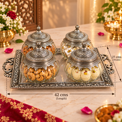 Four ornate silver-toned glass jars with matching lids are arranged on a decorative silver tray. The jars contain nuts and sweets, and the scene is set on a marble table with floral decorations and lit candles.