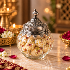 A clear glass jar with an ornate silver-toned lid, filled with round beige sweets and rose-petal pieces, set on a marble surface with warm decorative lights and flowers in the background.