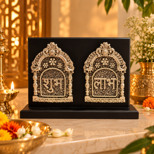 A black rectangular decorative box or tray holding two ornate silver-toned arched panels with Devanagari script and floral detailing, displayed on a marble surface with warm ambient lighting and floral props around it.