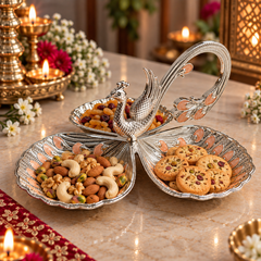 A decorative silver peacock-shaped serving tray with three petal-like compartments filled with nuts, dried fruits, and cookies, displayed on a marble table with lit candles and floral decor in the background.
