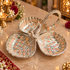 A decorative silver-toned metal tray shaped like a peacock with four leaf-like petals and a curved handle, displayed on a beige marble surface with warm ambient lights and floral decor in the background.