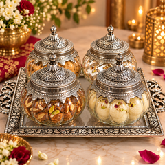 Four ornate silver-toned glass jars with decorative lids, arranged on a matching silver tray. The jars contain traditional sweets, including one filled with almonds and one with cream-colored pastries.