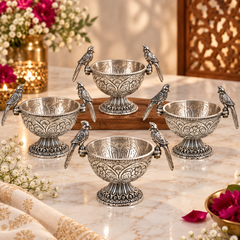 Five ornate silver pedestal bowls with bird-shaped handles, arranged on a marble tabletop with decorative flowers and a wooden tray in the background.