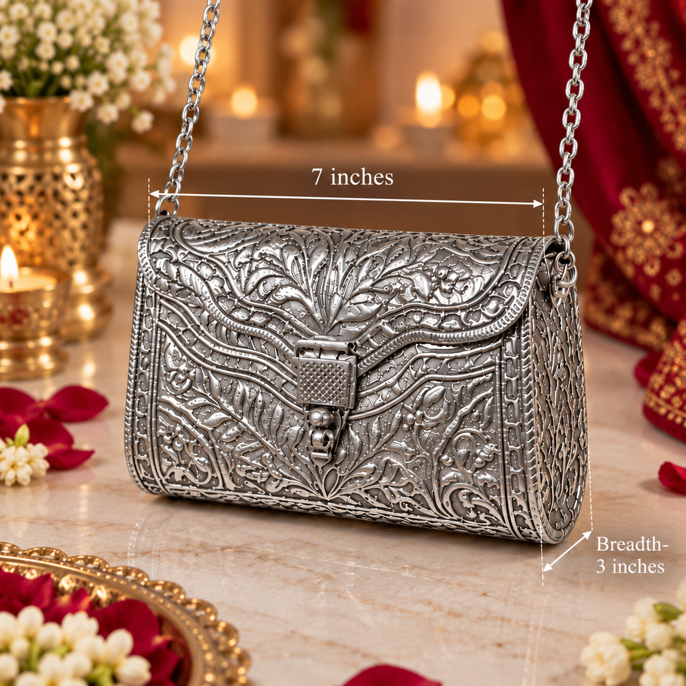 An ornate silver embossed handbag with a chain strap, shown on a marble surface with decorative flowers, candles, and red fabric in the background. White measurement lines and text indicate dimensions: 7 inches wide and 3 inches deep.