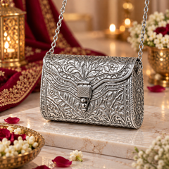 An ornate silver embossed handbag with a chain strap, displayed on a marble surface with red fabric, lanterns, and flowers in the background.