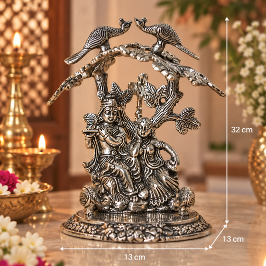 An ornate silver-toned metal sculpture of a seated couple under a tree with two peacocks on a branch, shown on a marble surface with decorative candles and flowers in a warm indoor setting. White dimension lines indicate 32 cm height, 13 cm width, and 13 cm depth.