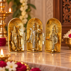 Three gold-toned Hindu deity figurines displayed side by side in arched niches, set on a polished marble surface with warm decorative background elements including flowers and lit candles.