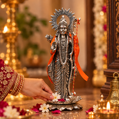 A detailed silver-toned Lord Hanuman statue on a decorative base, adorned with an orange ribbon, set in a warm, decorated home altar environment with a hand placing white flowers near it.