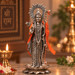 A detailed silver-toned metal statue of Lord Hanuman standing on a lotus base, draped with an orange cloth, displayed on a marble surface with lit diya candles and a warm, softly blurred background.
