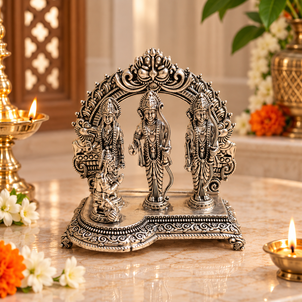 An ornate silver-toned Hindu deity idol with three standing figures under an arched decorative canopy, displayed on a marble surface with lit oil lamps and flowers in a warm indoor setting.