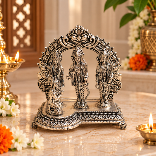 An ornate silver-toned Hindu deity idol with three standing figures under an arched decorative canopy, displayed on a marble surface with lit oil lamps and flowers in a warm indoor setting.