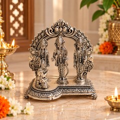 An ornate silver-toned Hindu deity idol with three standing figures under an arched decorative canopy, displayed on a marble surface with lit oil lamps and flowers in a warm indoor setting.
