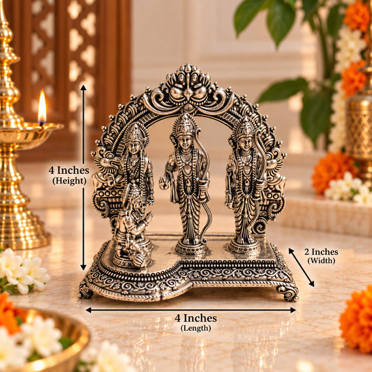 An ornate silver-toned Hindu deity figurine set on a decorative base, shown with dimension arrows and labels indicating 4 inches in height, 4 inches in length, and 2 inches in width. The scene is styled with a lit brass oil lamp, flowers, and greenery in a warm indoor setting.